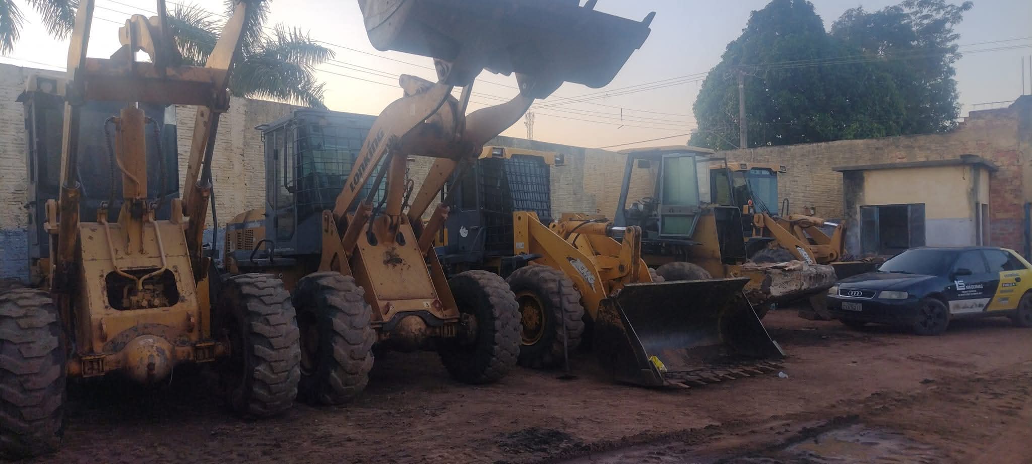 pátio com várias retroescavadeiras e máquinas pesadas amarelas estacionadas em terreno de terra, com um carro 
