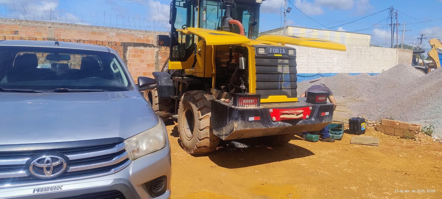 Caminhonete Toyota Hilux cinza estacionada ao lado de uma pá-carregadeira amarela Forza em manutenção por um m