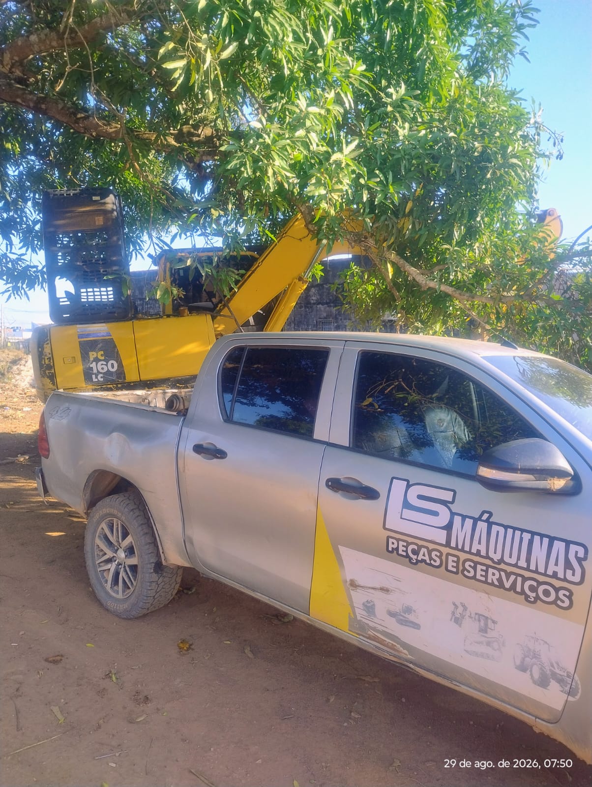 Caminhonete prata com adesivo LS Máquinas Peças e Serviços estacionada sob uma árvore, com escavadeira amarela