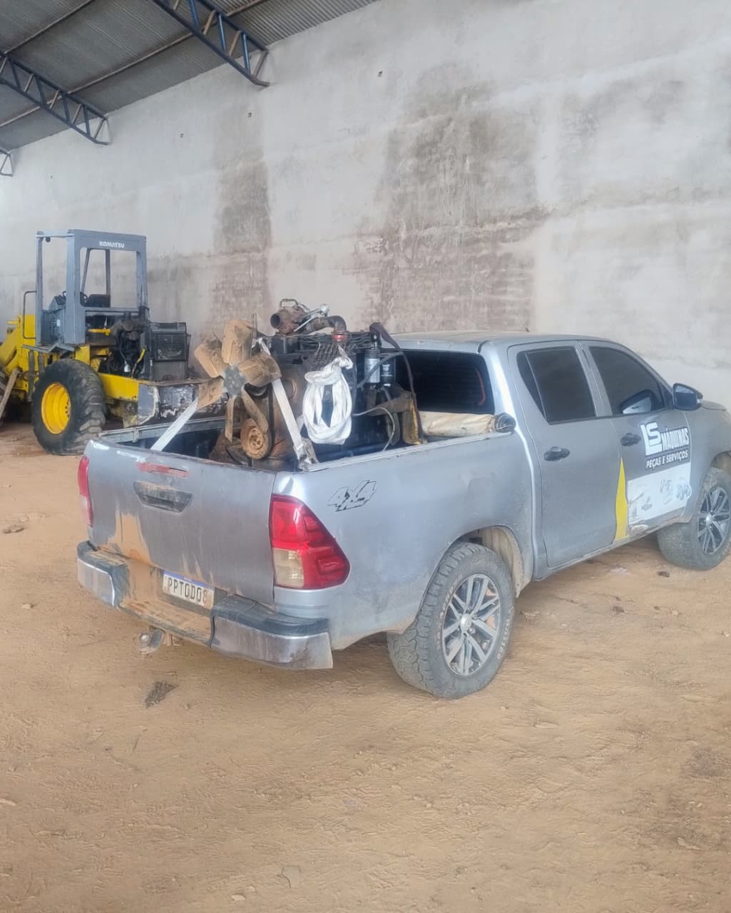 picape prata carregada com peças de motor em galpão de terra, com uma pá carregadeira Komatsu amarela ao fundo