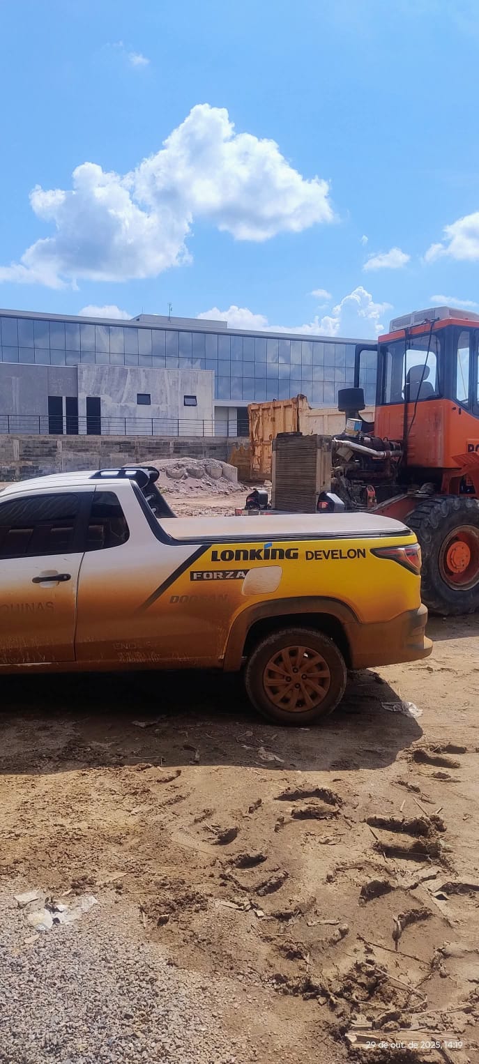 picape amarela e branca com grafia Lonking Develon estacionada em terreno de terra ao lado de uma pá carregade